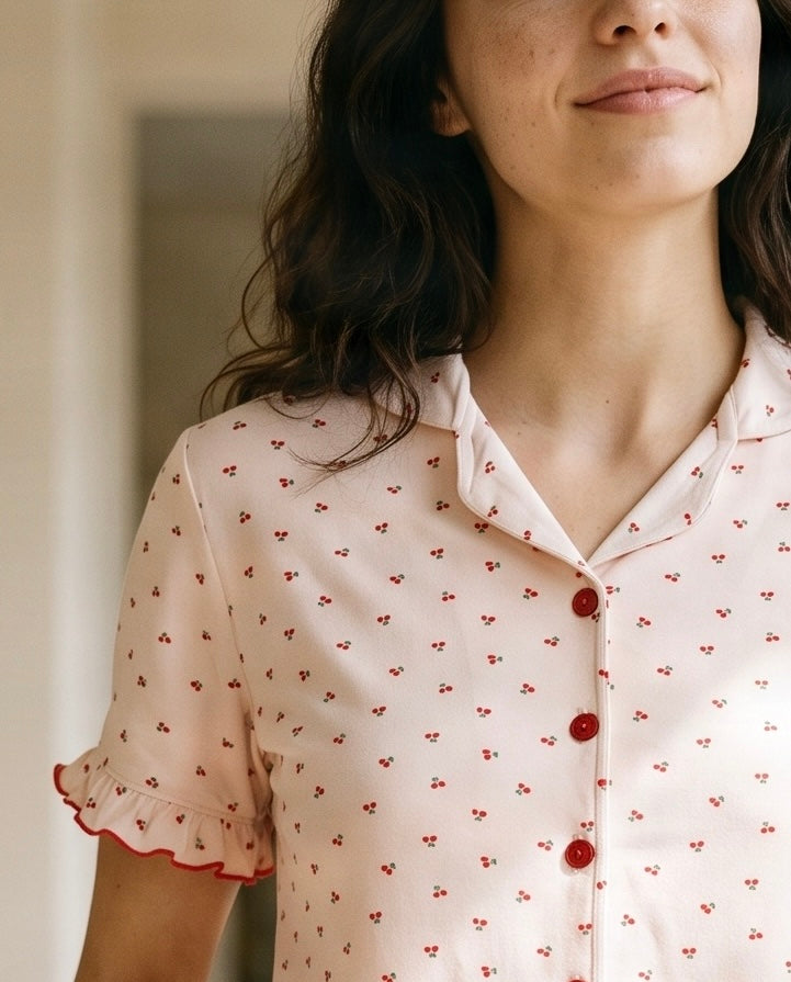 A close-up of a woman wearing a light pink short-sleeve button-up shirt with a small red cherry print and red ruffled trim on the sleeve cuffs.