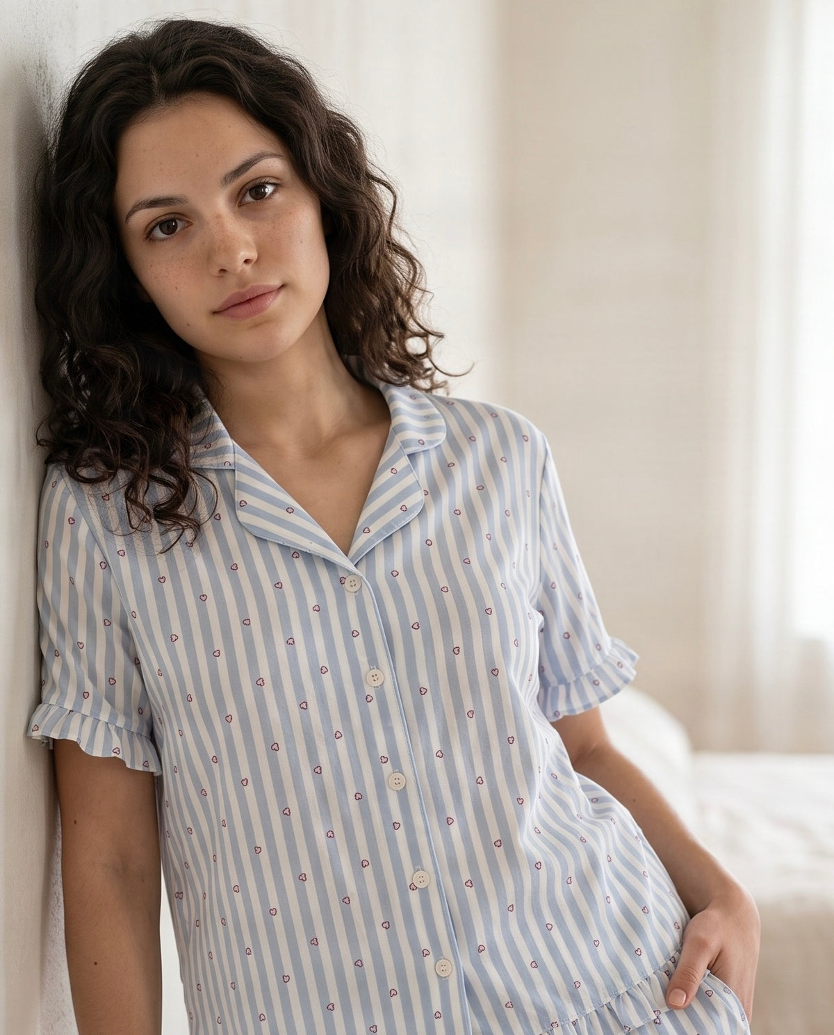 A woman wearing a light blue and white striped short-sleeve pajama shirt with a small floral pattern, leaning against a neutral wall in a softly lit room.
