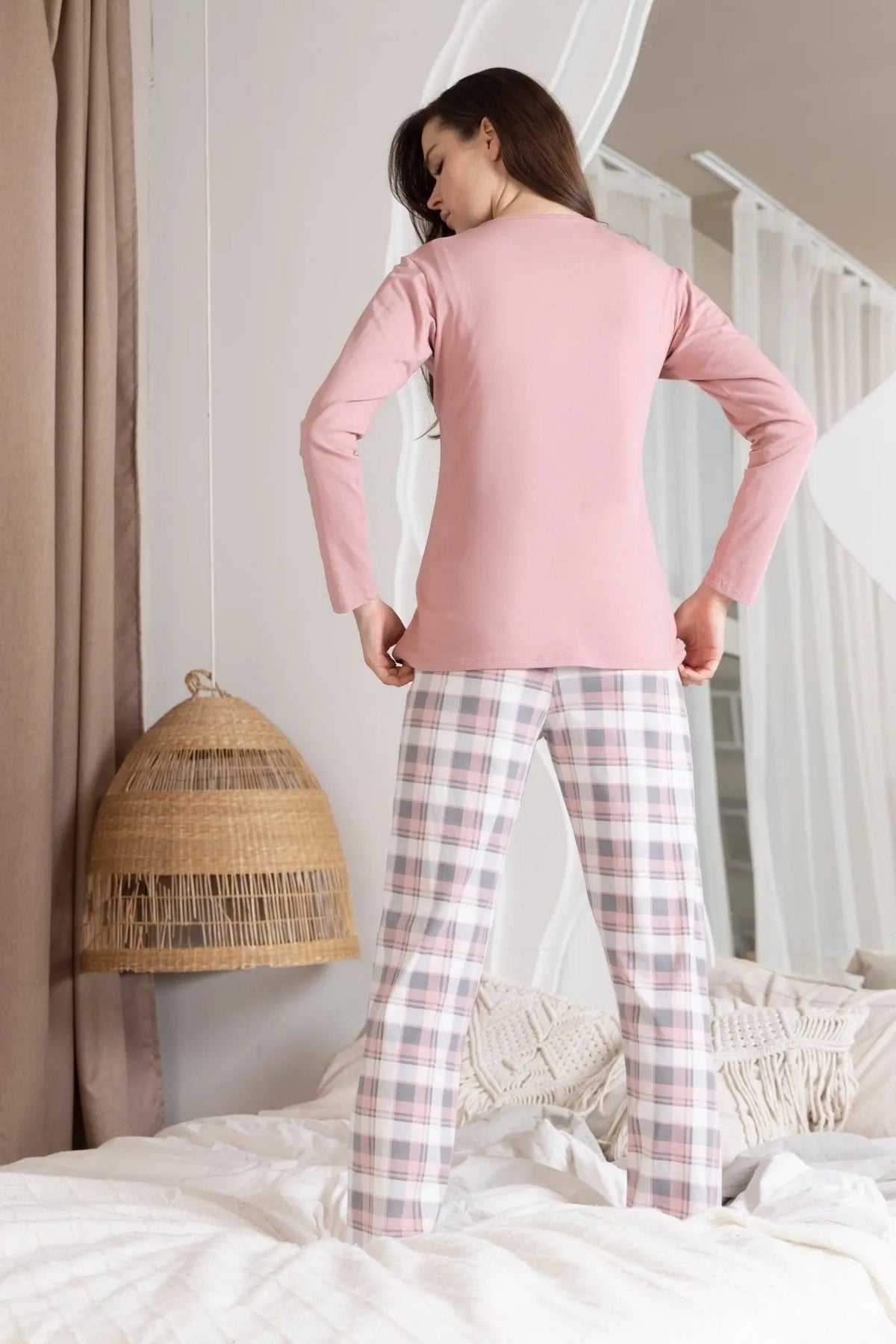 Woman wearing pink pajama top and plaid pants standing in a bedroom.
