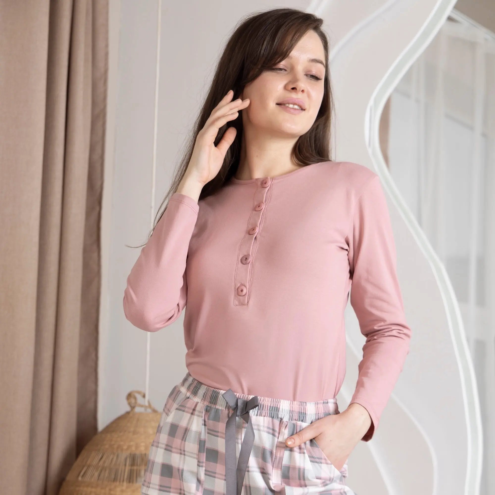 Woman wearing a pink long-sleeve top and plaid pants in a home setting.