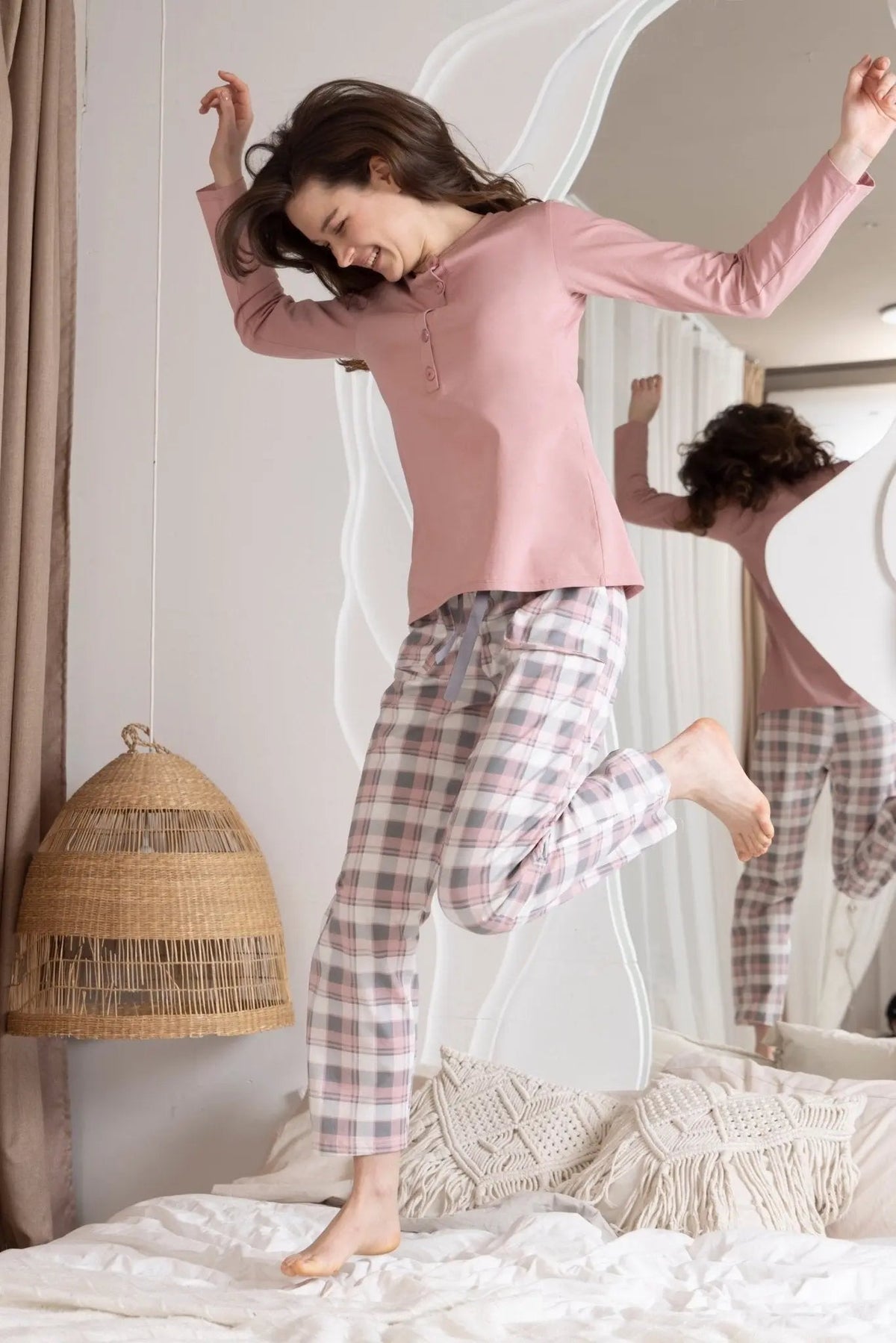 Woman in pink top and plaid pants standing on a bed in a bedroom.