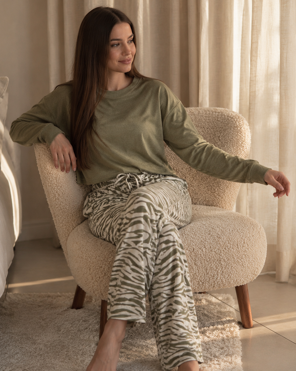 A woman sits barefoot in a cream boucle armchair in a softly lit room, wearing an olive green long-sleeve top and beige-and-white zebra-print pajama pants.