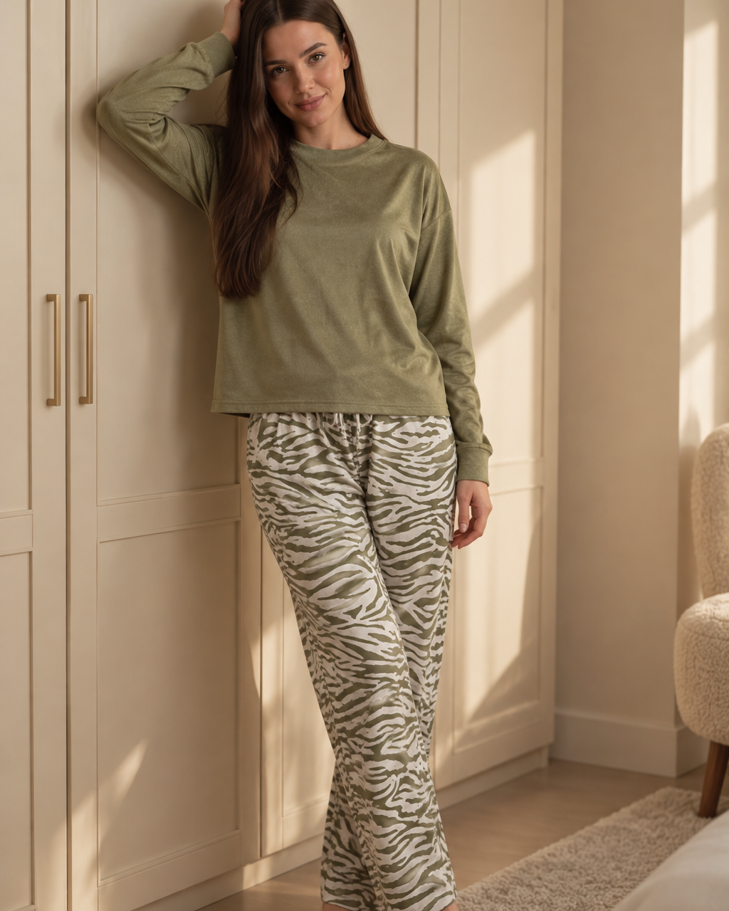 A woman wearing a two-piece pajama set consisting of an olive green long-sleeve top and beige-and-green zebra-print pants, standing barefoot in a brightly lit bedroom next to a cream-colored wardrobe.